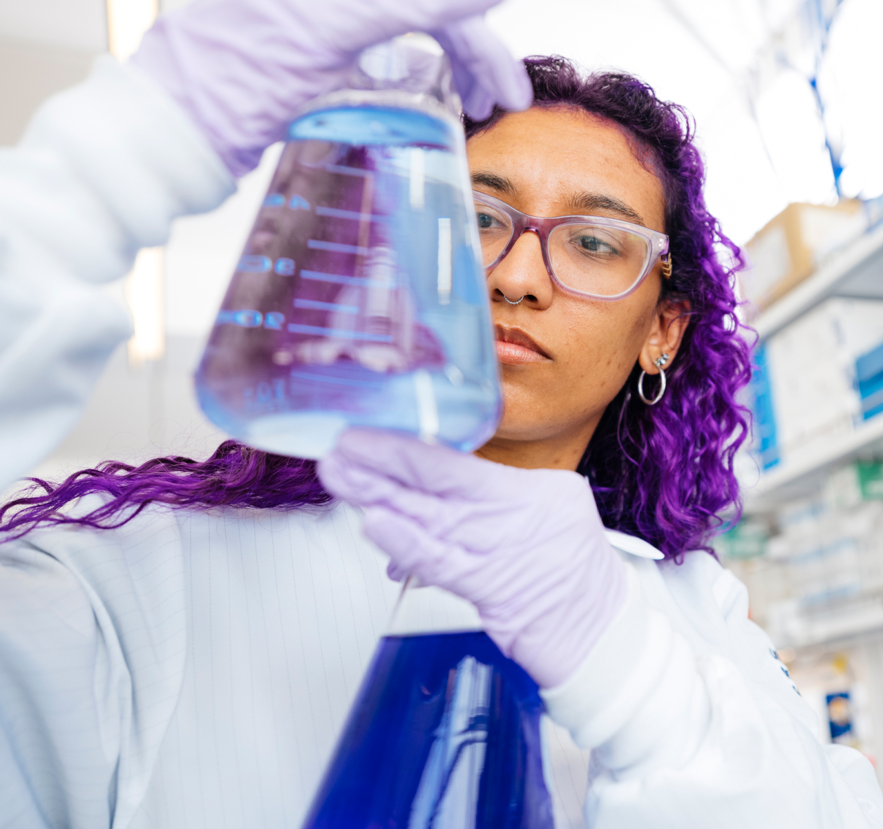 A student scientist with purple hair and glasses measures liquids in a couple Erlenmeyer flasks