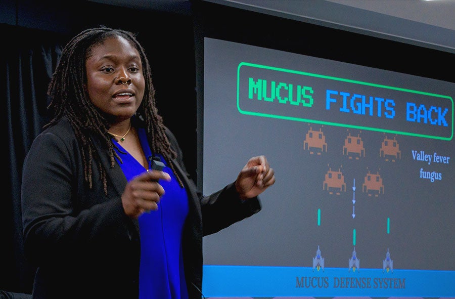 Tahirah Williams presenting her first-place winning talk, standing in front of her slide entitled 'Mucus Fights Back' which uses Space Invaders imagery to illustrate how our mucus defense system can defend against Valley Fever fungus.